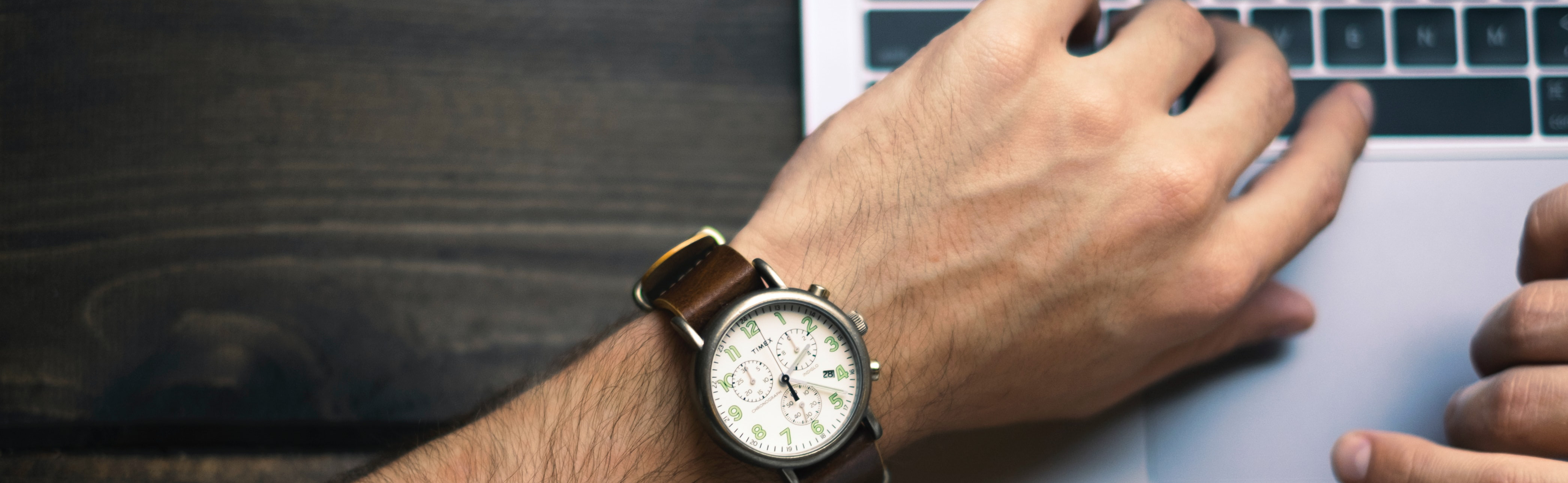 A hand with an analog wrist watch hovering over a laptop keyboard